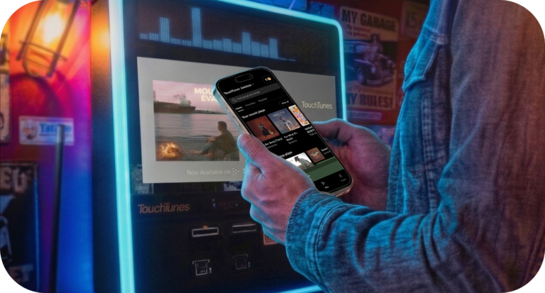 A person uses a smartphone app to choose songs on a TouchTunes digital jukebox in a colorful bar setting, with the jukebox screen glowing and displaying music content.