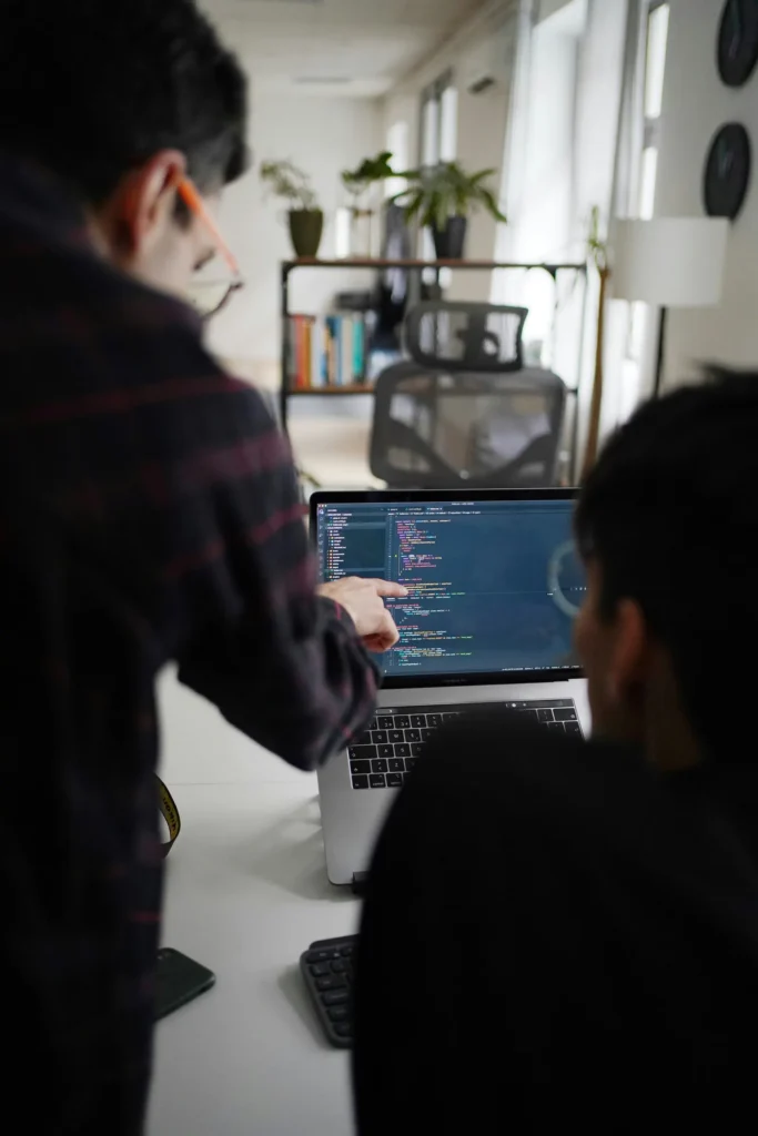 Developer pointing at code on a laptop screen while a colleague reviews it, illustrating what vibe coding looks like when AI-generated code gets a human review