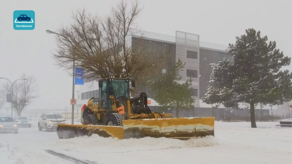 Wheel loader with a snow plow blade clearing a snowy street during a snowfall, with the Terrebonne parking app icon in the corner