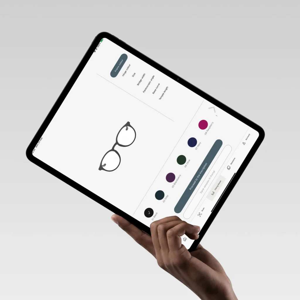 Hand holding an iPad with the YOU MAWO Fitting Room, showing frame colours and fit settings such as bridge width